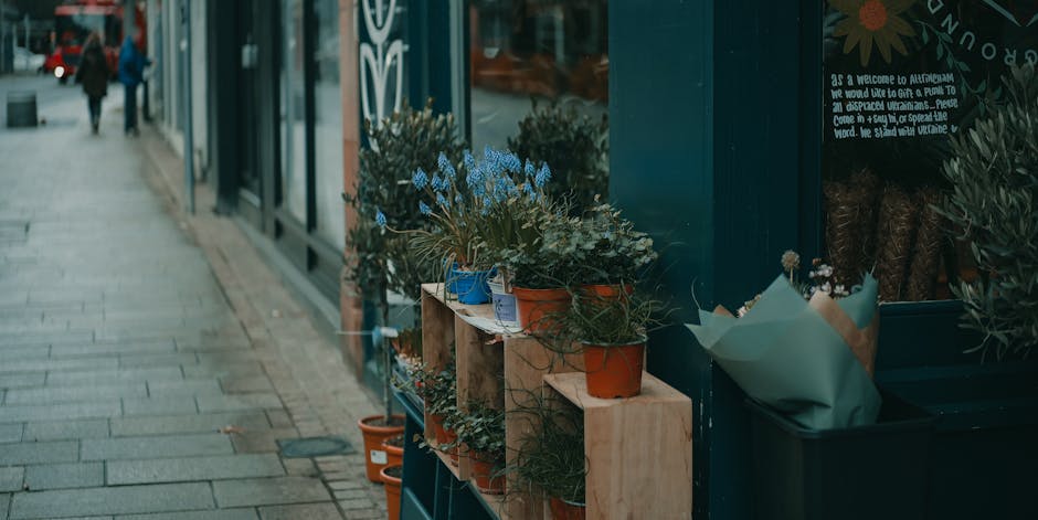 A display of potted plants and floral arrangements placed on wooden shelves outside a shop, featuring a variety of greenery and small flowering plants with blue, orange, and natural-colored pots. The