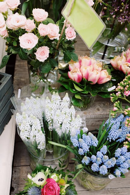 A variety of fresh flowers displayed on a wooden surface, including a bouquet of pale pink roses with lush green leaves, arranged in a tall clear glass vase; a separate arrangement of soft pink tulips